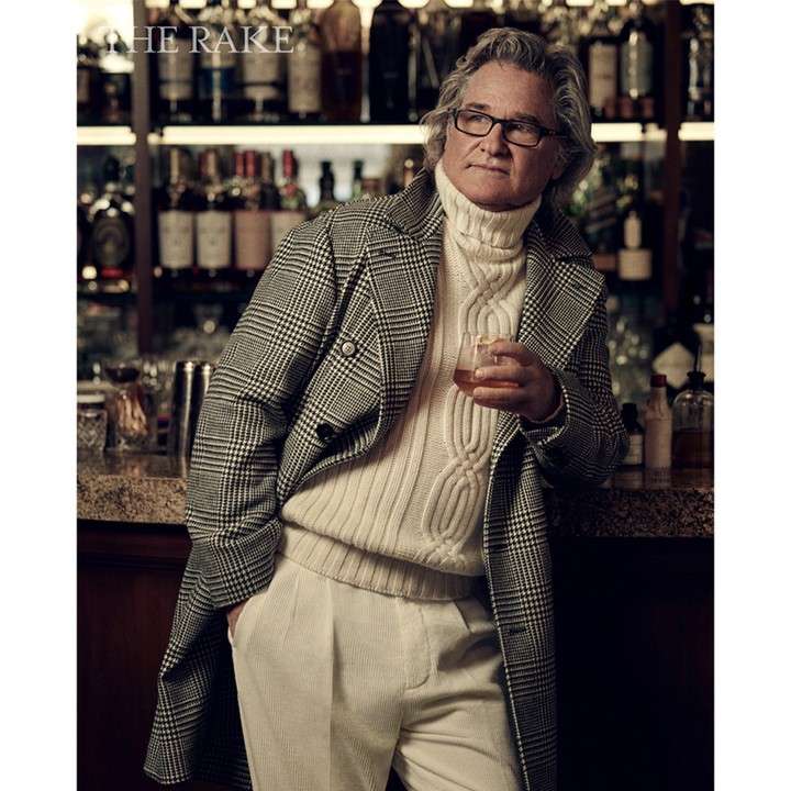 Adult male poses indoors in a glen plaid overcoat and turtleneck sweater while holding a glass of amber liquid near a bar.