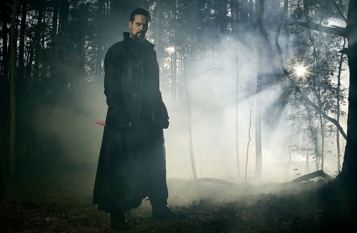 Man in long black coat standing in forest, holding knife, foggy, cinematic, dramatic lighting.