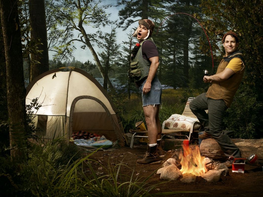 Two men in casual outdoor wear camping, one is fishing by the campfire near a tent surrounded by a forest-like background in a fun, playful setting.