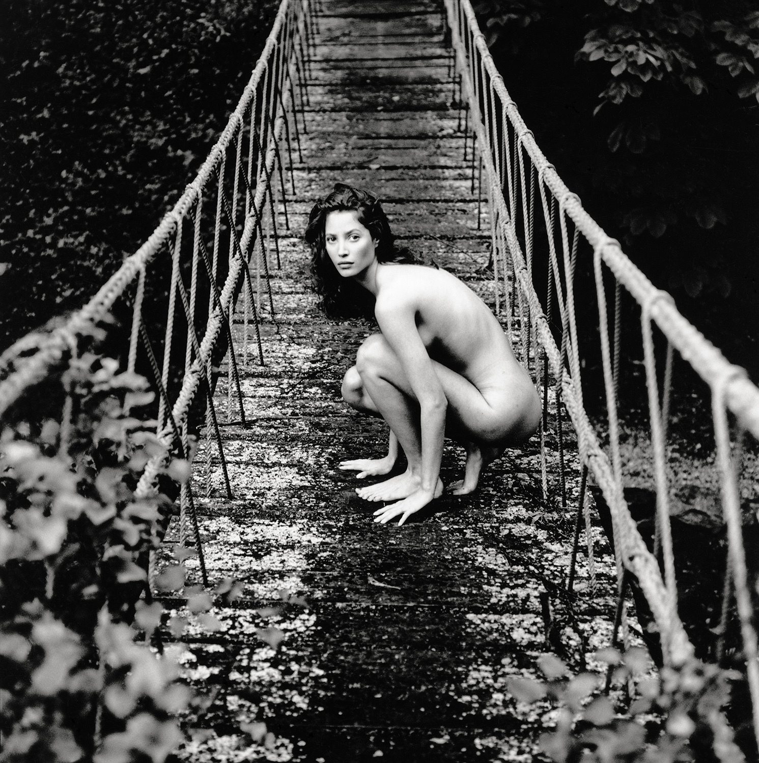 Nude adult female crouches on rope bridge, gazing at the camera in a black and white photograph, surrounded by dense foliage.