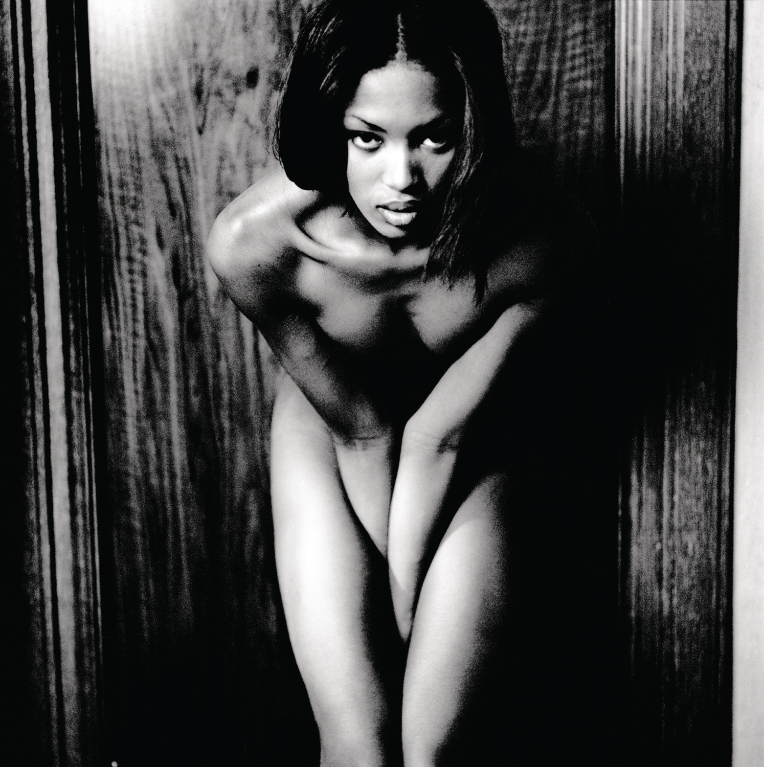 Black and white portrait of Naomi Campbell leaning forward against a dark wooden door frame.