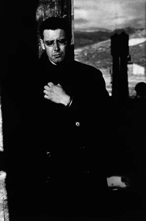 Monochrome portrait of a man in glasses and a dark coat standing outdoors in front of a hilly landscape by Anton Corbijn.