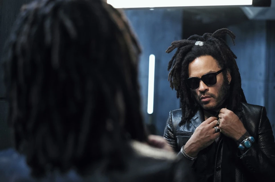 Portrait of an adult male with dark hair and sunglasses, wearing a black jacket and jewelry, reflected in a mirror with cool lighting.