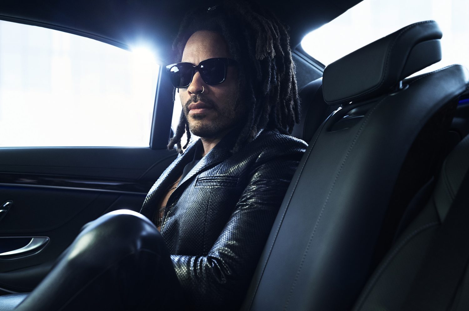 Adult male with dreadlocks and sunglasses sits in the back seat of a car wearing a black jacket; interior shot, automotive theme.