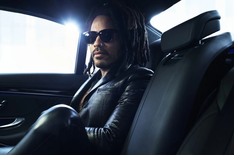 Adult male with dreadlocks and sunglasses sits in the back seat of a car wearing a black jacket; interior shot, automotive theme.