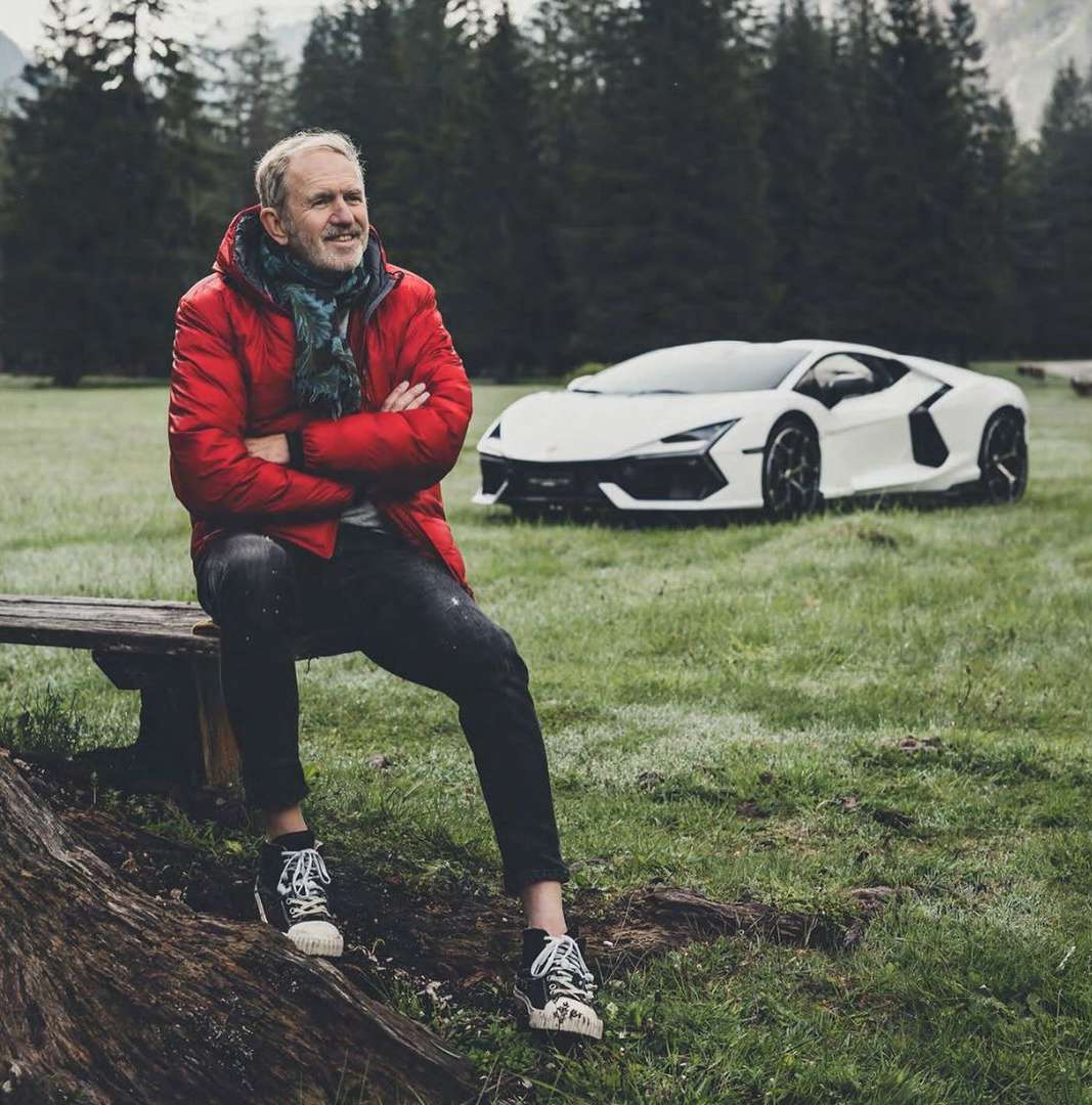 Adult man sits on a wood bench near a white Lamborghini in a grassy field with trees in the background. The man is smiling.