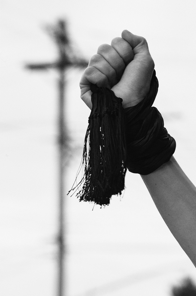 Monochrome shot of a raised fist holding black tasseled cloth in an outdoor setting with an out-of-focus utility pole in the background.