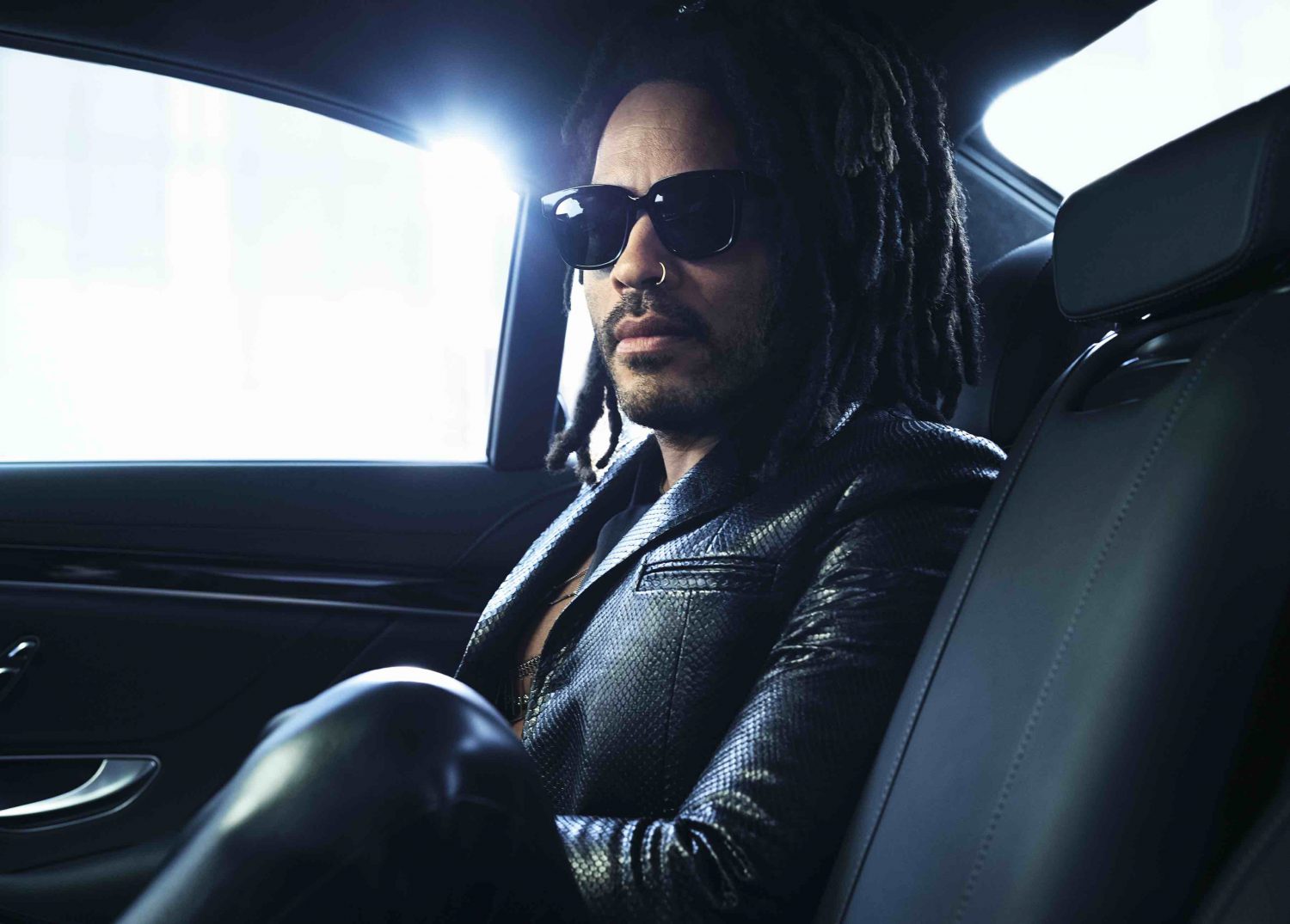 Stylish man with dreadlocks and sunglasses sits inside a car, wearing a black leather jacket, in an indoor, automotive setting.