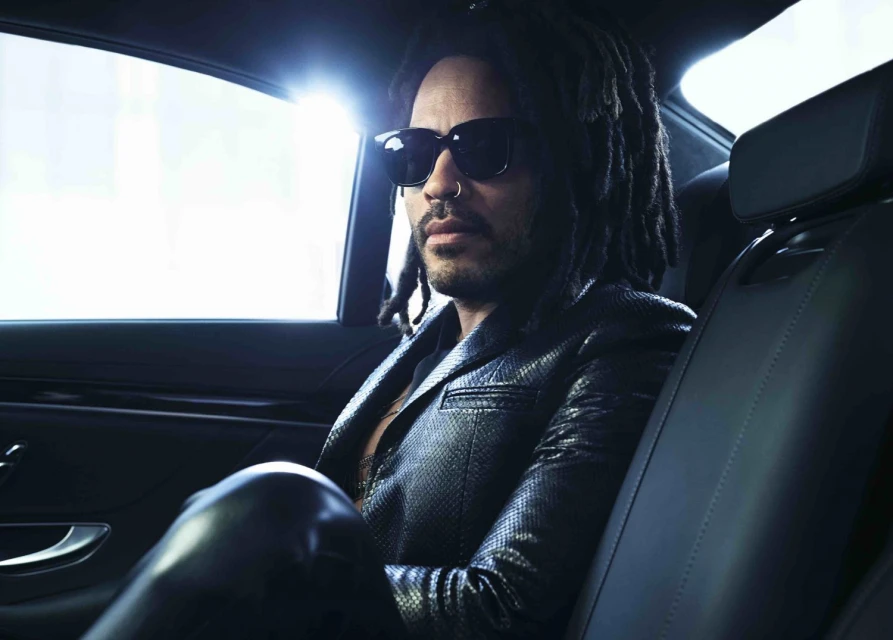 Stylish man with dreadlocks and sunglasses sits inside a car, wearing a black leather jacket, in an indoor, automotive setting.