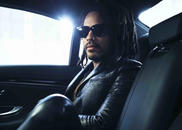 Stylish man with dreadlocks and sunglasses sits inside a car, wearing a black leather jacket, in an indoor, automotive setting.