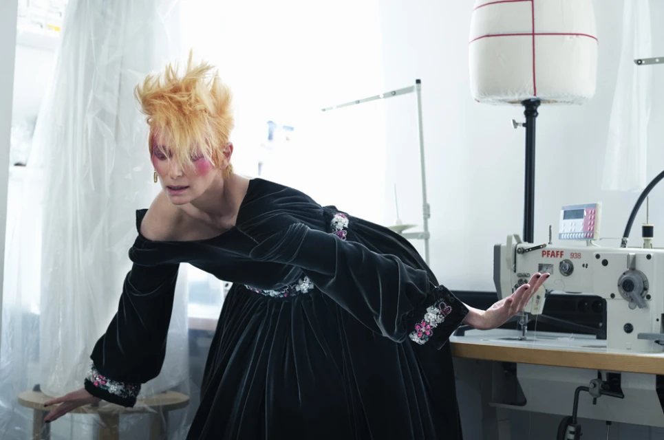 Woman in a dark off-shoulder dress, posing in a sewing room with a dress form and sewing machine. Bright, creative fashion studio shot.
