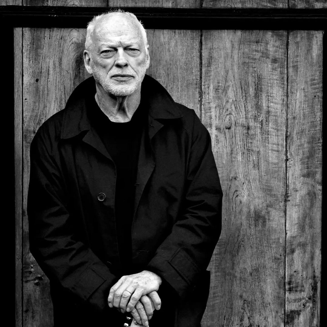 Black and white portrait of an older man with short white hair, wearing a black coat, standing in front of a wooden door.