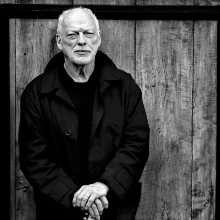Black and white portrait of an older man with short white hair, wearing a black coat, standing in front of a wooden door.