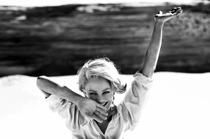Black and white portrait of a smiling woman raising one arm and covering her mouth with her hand, wearing a light shirt.