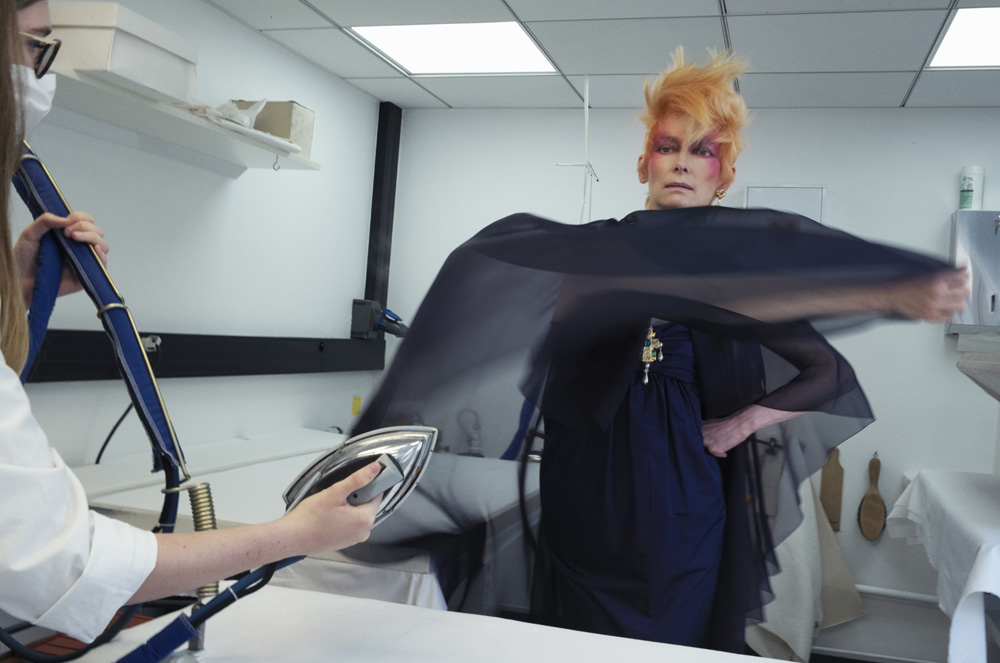 Adult female with spiky orange hair poses as a second person uses a steam iron to smooth a black fabric, indoors in a white room.