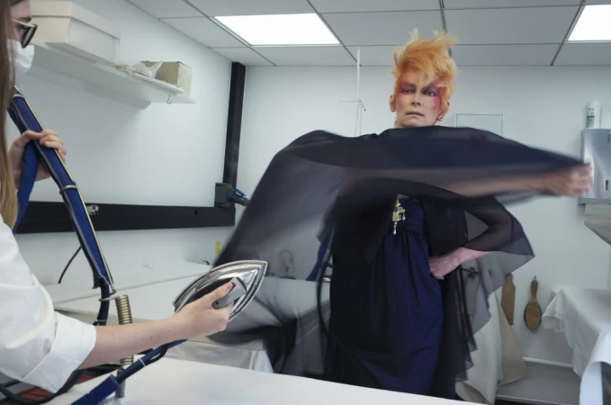 Adult female with spiky orange hair poses as a second person uses a steam iron to smooth a black fabric, indoors in a white room.