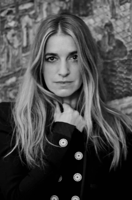 Black-and-white portrait of a woman with long blond hair, wearing a dark jacket with decorative buttons, set against an ornate wall.