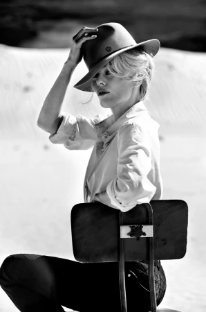 Black and white portrait of a woman wearing a fedora hat and white shirt, seated backwards on a chair outdoors against a sandy dune background.