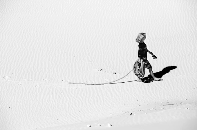 Woman walking barefoot across sand dunes, dragging a coiled rope behind her in a high-contrast monochrome photograph.