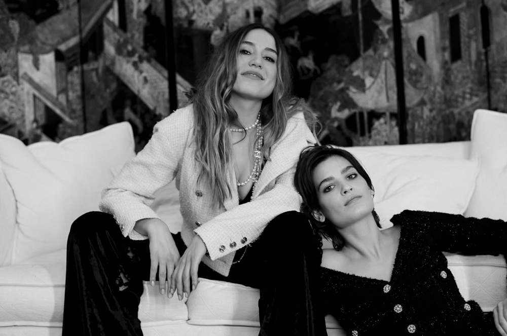 Two women pose on a white couch in front of a painterly backdrop, wearing fashionable tweed jackets and black pants, black and white.