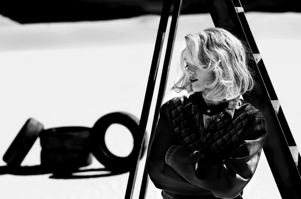 Blonde woman poses with folded arms in black and white, set against tires and a ladder, creating a minimalist, edgy fashion statement.