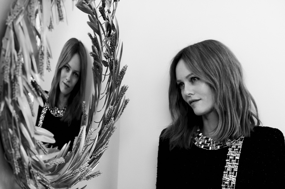 Portrait of Stella McCartney wearing a dark embellished top, captured in monochrome with her reflection in a decorated circular mirror.