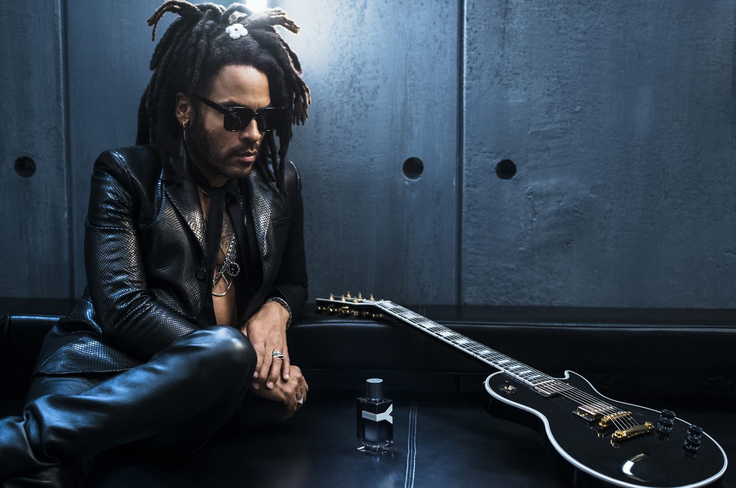 Lenny Kravitz seated with a guitar and fragrance bottle in a sleek studio, showcasing a blend of music and modern elegance.