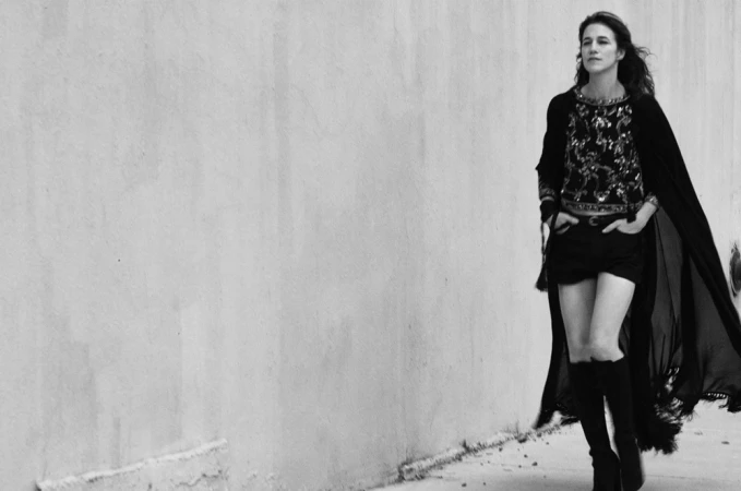 Black and white portrait of a woman walking alongside a plain light wall wearing tall boots and a patterned top