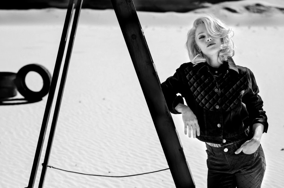 Monochrome shot of a blonde woman in a quilted jacket and patterned pants, leaning on a wooden structure in a sandy, desert landscape.