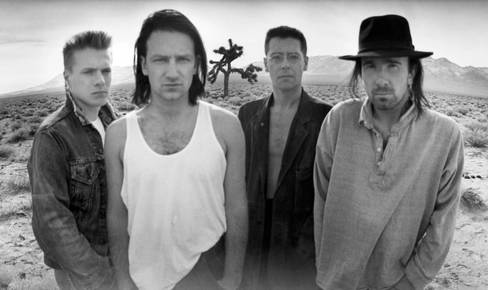 Black and white portrait of the band U2 standing in a desert landscape, featuring Bono, The Edge, Adam Clayton, and Larry Mullen Jr.