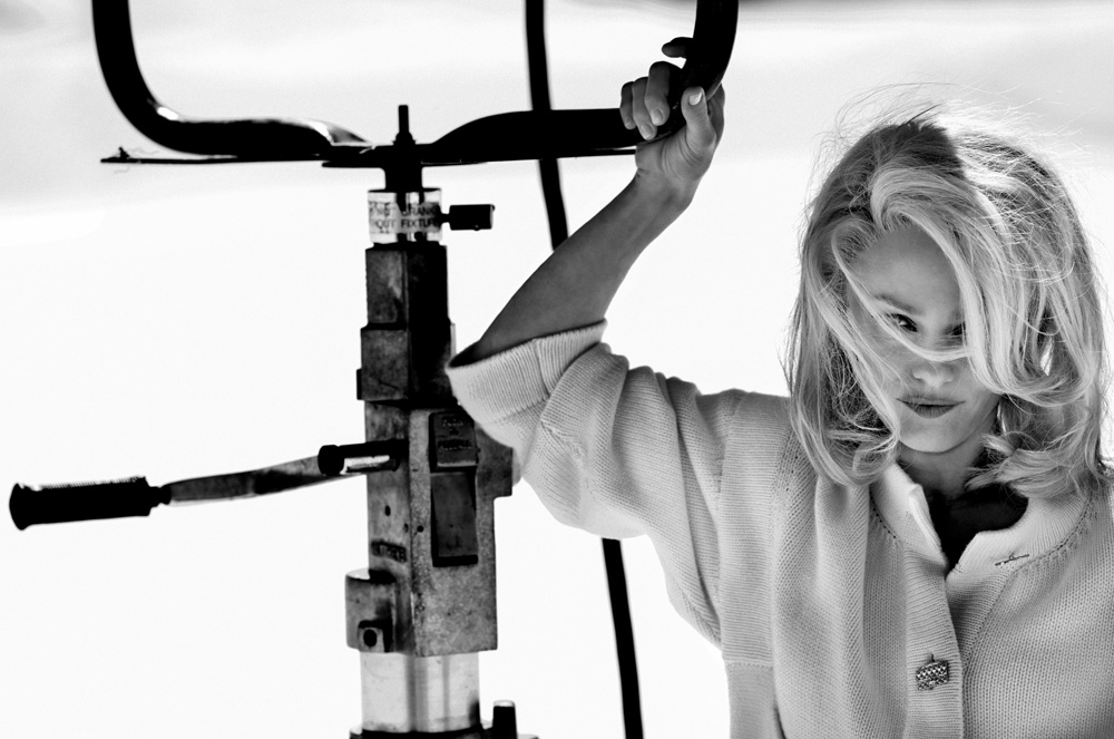 Monochrome portrait of a blonde woman in a white cardigan holding onto an industrial metal structure against a bright background.