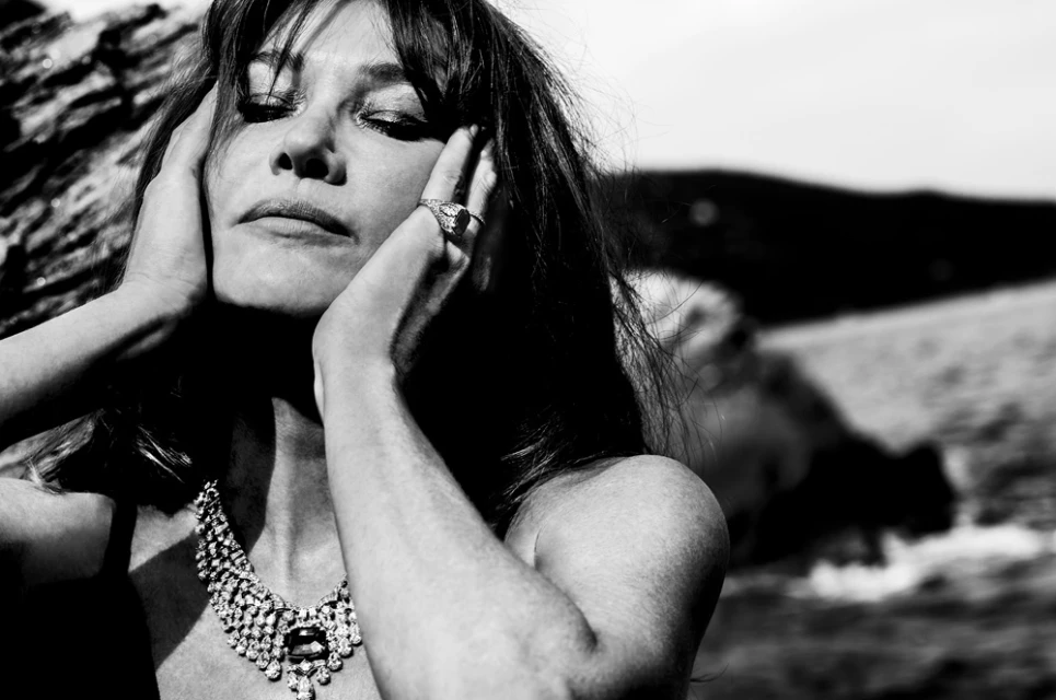 Black and white portrait of an adult female with dark hair wearing a necklace and ring, touching her face, with ocean in background.