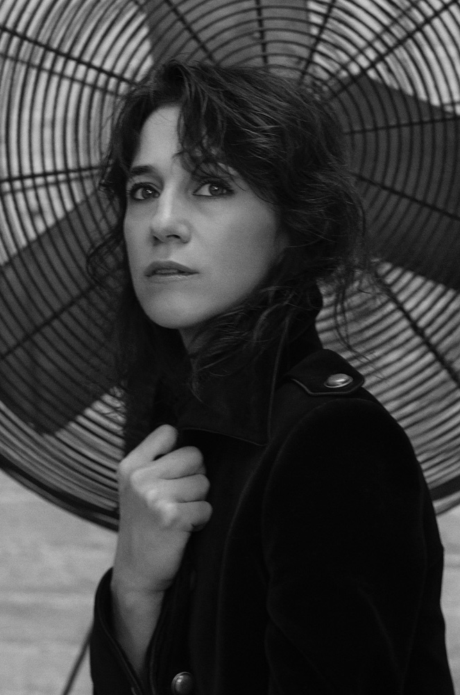 Monochrome portrait of a woman with dark hair wearing a dark coat, standing in front of a large vintage fan.