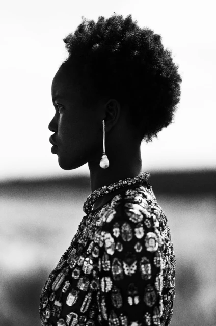 Profile portrait of a stylish Black woman in a beaded jacket and dangling pearl earring, set against an out-of-focus outdoor background.