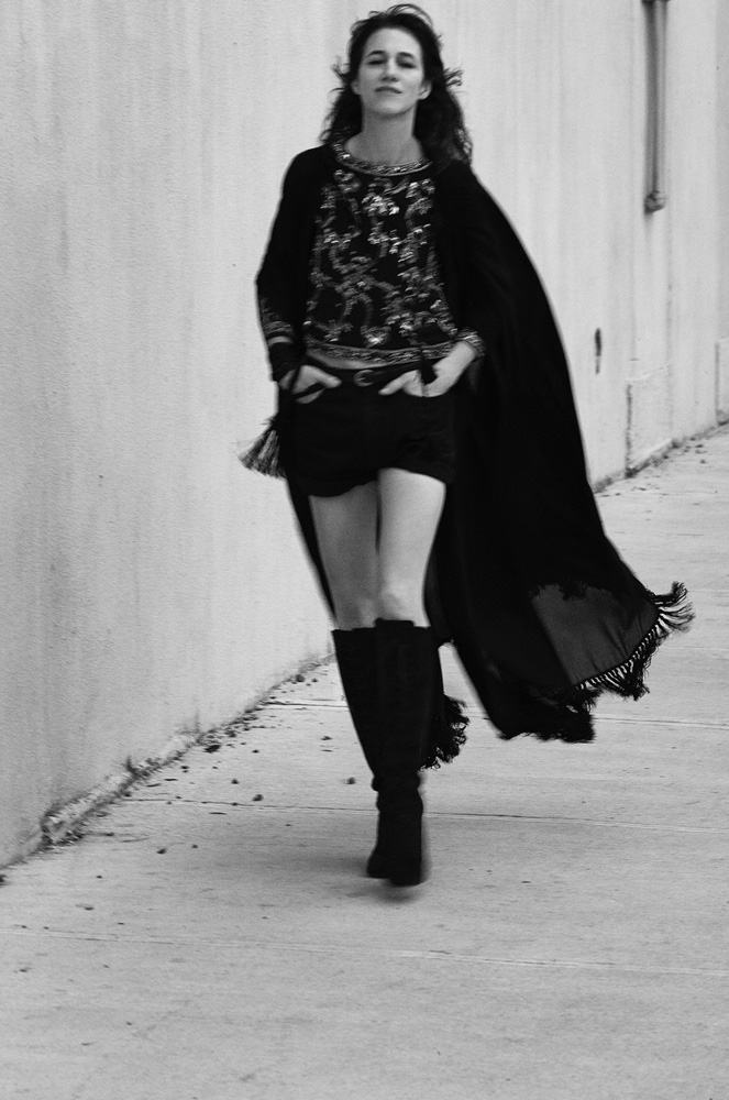 Monochrome portrait of a woman walking down a sidewalk wearing a dark cape and tall boots.
