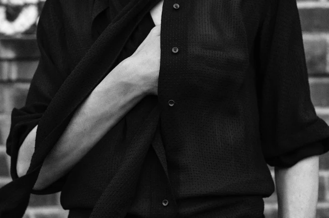 Close-up of a person wearing a textured dark buttoned shirt with hand placed inside the opening, photographed by Anton Corbijn.