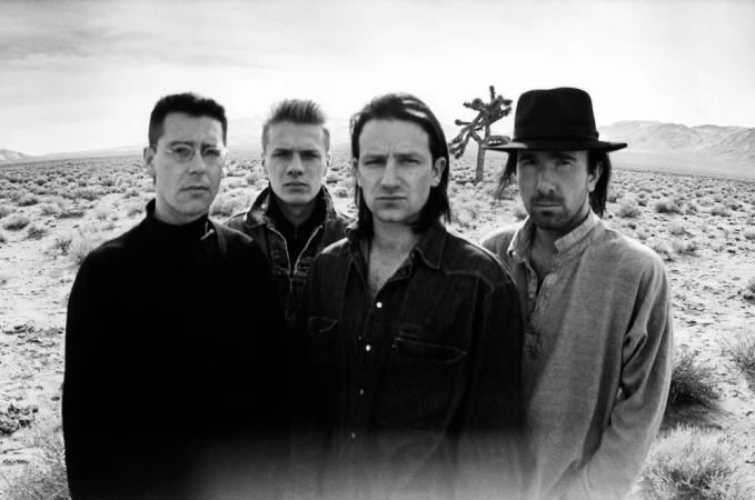 Black and white shot of four adult men in casual clothing standing in a desert landscape in front of a metal sculpture.