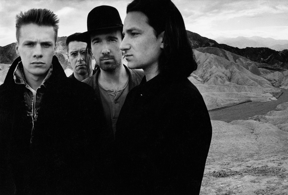 Black and white portrait of U2 band members outdoors with mountains in the background; Bono stands in front wearing a jacket and hat.