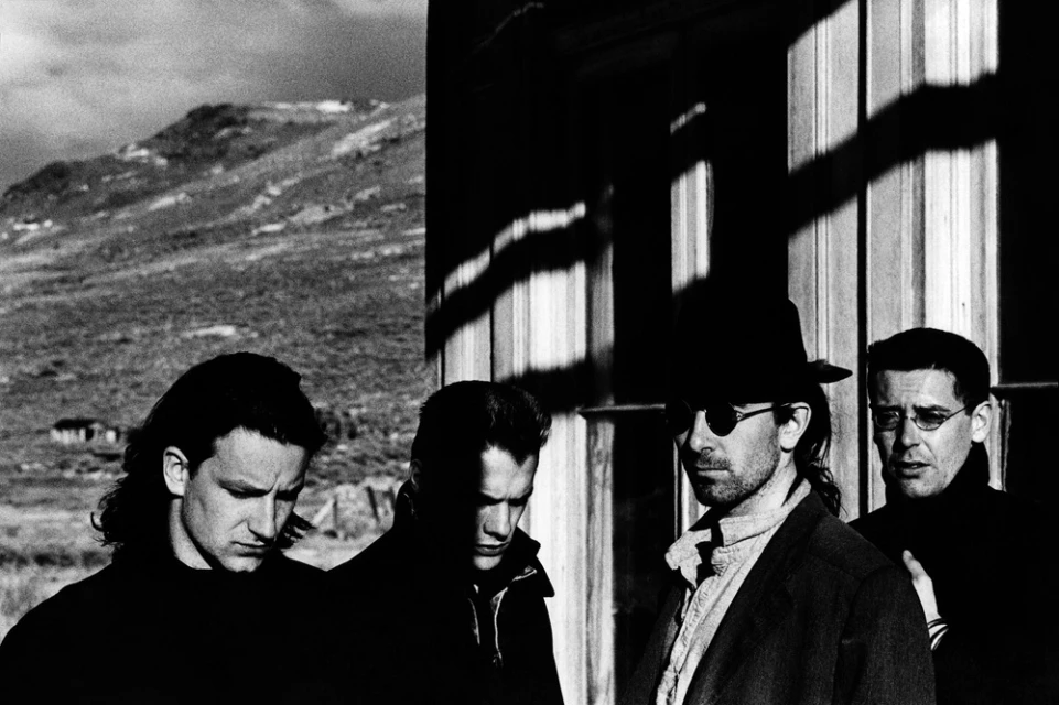 Black and white portrait of the band U2, standing outside against a mountainous background and in front of a building with windows.