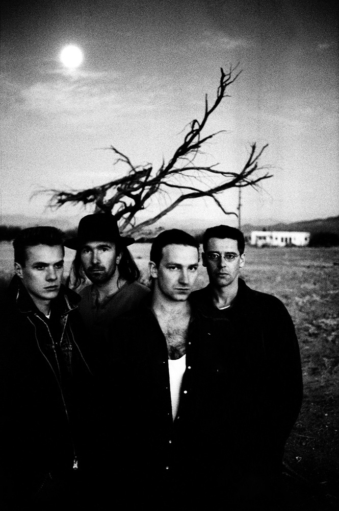 Black-and-white image of a music band of four men in a field at dusk, with a dead tree and a building in the background.