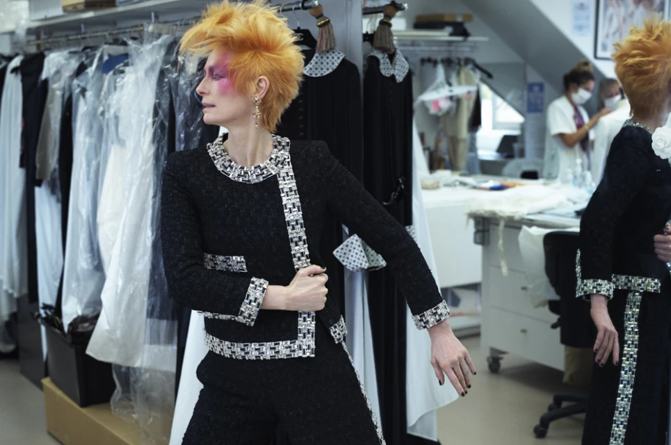 Adult woman poses in a black tweed suit in a fashion studio with garments visible on racks in the background. Professional fashion shot.