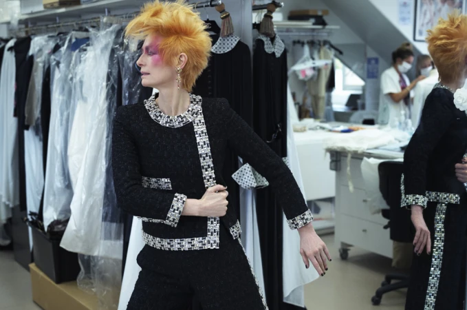 Adult woman poses in a black tweed suit in a fashion studio with garments visible on racks in the background. Professional fashion shot.
