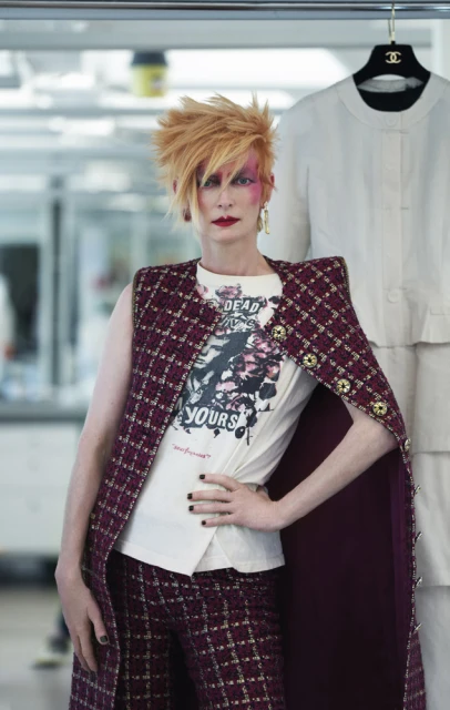 Adult female fashion model with red hair wearing tweed vest and pantsuit, standing in a studio environment, posing with Chanel clothing.