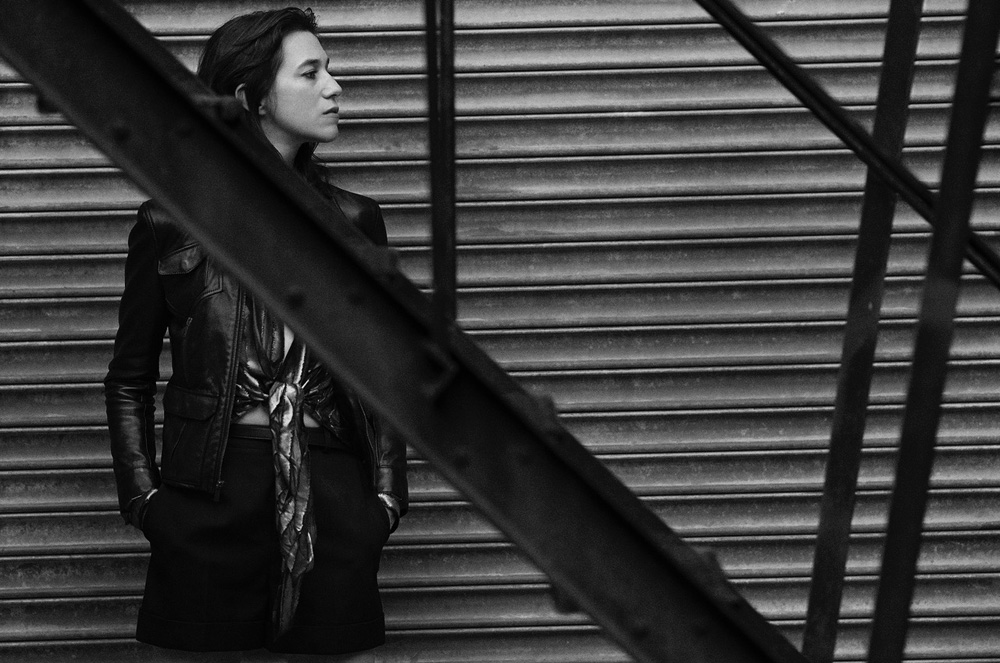Adult woman with hands in pockets poses in leather jacket in front of a metal garage door in a black and white portrait.