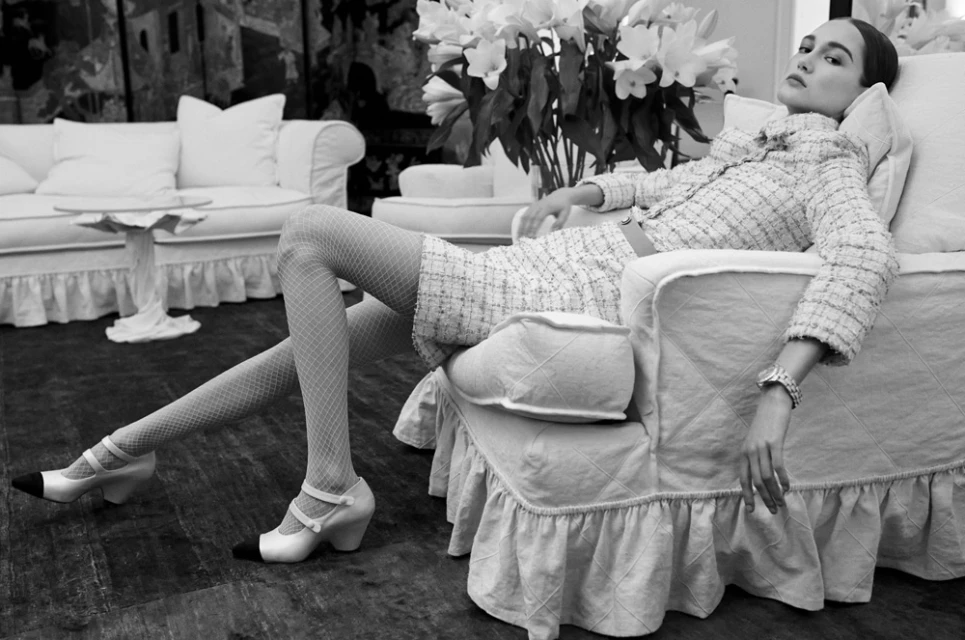Elegant woman in tweed suit reclines on white vintage armchair in monochrome, flowers and white furniture in soft focus background.