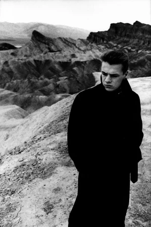 Monochromatic portrait of a man in a coat outdoors. Landscape includes mountains and exposed rock formations, creating a stark contrast.