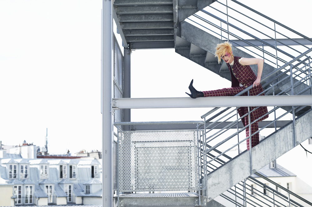 A model in a checkered suit strikes a pose on a metal staircase against a backdrop of Parisian rooftops and bright sky.