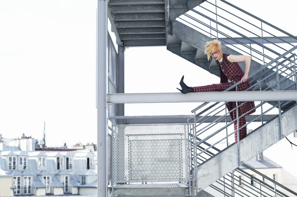 A model in a checkered suit strikes a pose on a metal staircase against a backdrop of Parisian rooftops and bright sky.