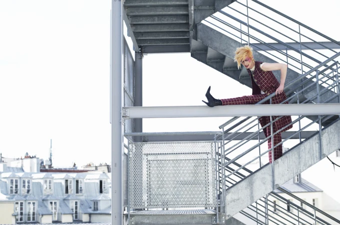 A model in a checkered suit strikes a pose on a metal staircase against a backdrop of Parisian rooftops and bright sky.