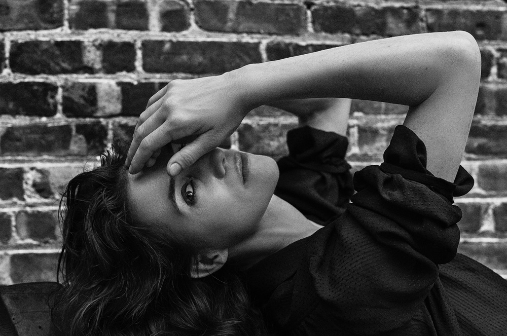 Black and white portrait of a woman reclining against a brick wall, covering her eyes with her hand in an outdoor setting.